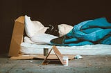 A makeshift bed on a concrete floor, comprising a white mattress and pillow, a blue blanket and large pieces of cardboard.