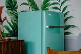 teal green refridgerator in a kitchen. A chair is in front of it, and painted leaves are behind it on the white wall.