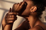 A brown-skinned Black woman with rings on her fingers. Her hands hide her face. She angles her body to the side.