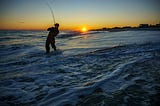 a person standing in the waves at sunset holding a fishing line