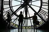 giant wall clock with 3 figures looking up to signify TIME