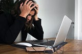 Image of woman in distress, gripping her head with her hands while staring at a laptop. Her glasses and phone are in the foreground next to her computer