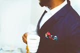 Black man with a beard in an expensive dark blue suit and crisp white shirt holding a cup of coffee.