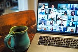 A mug of coffee next to a MacBook; the Macbook has a Zoom call ongoing.