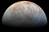 The surface of Europa against the blackness of space. The surface is light blue and white with reddish cracking all across it.