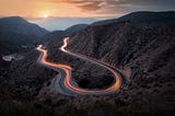 Curvy road surrounded by mountains with sunset over water on the horizon