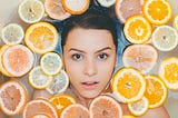 A make-up free woman looks out from a bathtub filled with sliced citrus fruit.