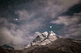 Night sky crowded with stars over a craggy and cloudy mountaintop