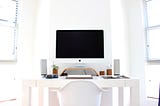 A neat white room with a picture of White Mac Monitor with symmetrically placed speakers, a white keyboard and a brown pot with green plant on one side and 2 brown desktop decorations of similar size on the other side of Monitor. All of this on a white table with a white ergonomic chair. A perfect symmetrical shot of this home office.