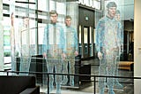 An artistic installation resembling the Star Trek transporter effect, featuring life-sized human figures seemingly formed from colorful beads suspended on vertical strings. The figures appear to be materializing or dematerializing in mid-air, capturing the iconic look of Star Trek’s transporter beam. The scene is set in a modern, spacious indoor area with natural light, glass walls, and minimalistic furnishings.