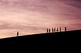 A group of people walking down a hill with a person leading them, there is a pink sunset in the background.