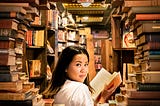 A woman reading a single paper book in the middle of piled books and books on shelves. She looks back at the viewer with a smile.