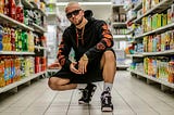 A tough guy wearing sunglasses and a hoodie crouching down in the drinks aisle of the supermarket