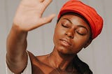 A young Black woman, wearing a white, flowy, one-shoulder, summer blouse and bright red-orange baker-boy hat, stands resolutely and peacefully with her eyes closed and her right arm extended out before her to bear an open palm that expresses the universal signal for the command “stop.”