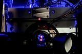 Close-up of a scientific instrument with wires and a circular metallic component, illuminated by blue light, featuring a small red dot at the center.
