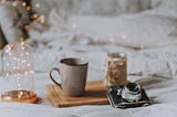 Book, open-faced on a bed with various items. A glittery light creates a magical feel next to a coffee mug on a tray and vintage camera.