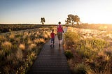 An image of a mother holding her son’s hand as they walk along a brightly lit path in the middle of a field.