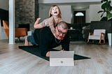 A multitasking father, doing a plank with his daughter sitting on his back, attempts to work on his computer.