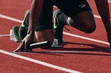 Relay racer at starting line, knee on the ground, toes set on blocks, holding a baton, ready to sprint. This image symbolizes the initial phase of market research, preparing businesses to launch forward with strategic insights.