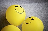 Three yellow smiley face balloons on a concrete grey floor.