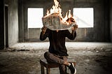 Man sitting on a stool inside an abandoned building holds a newspaper that is fire
