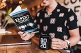 White guy with short sleeve shirt holding black mug with numbers on it reading a book about expert secrets