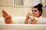 A woman in a bathtub eating a container of takeout noodles. Her hair is in a bun and her feet are elevated on the edge of the tub.