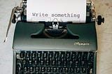 A vintage typewriter holds a piece of paper with the direct call to action: ‘Write something,’ representing the daily, focused work of a writer.