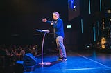 A man in casual dress addresses an audience from a stage