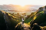 Woman standing on top of hill overlooking nature fields during sunset