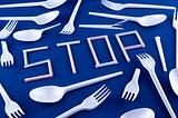 Bunch of single-use plastics spread out on a blue table, with the word “STOP” made from plastic straws, surrounded by many plastic spoons & forks.