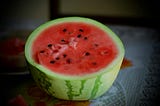 A picture of half of a watermelon, with its red center facing upwards on a tablecloth. The watermelon’s red center is also filled with dispersed black seeds.