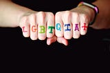 A photo of LGBTQIA+ written in rainbow-colored letters across someone’s knuckles