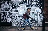 homme marchant avec son vélo, il téléphone. En arrière plan, fresque murale évoquant l’écosystème saturé de la ville.