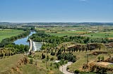 A photo of a landscape, with green fields, it’s a sunny day, a road zig-zags downhill and goes towards a bridge, that crosses the Duero river in Toro, Spain.
