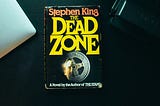 A book by Stephen King, Titled: The dead zone, on a background with a macbook laptop peaking out.