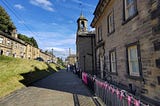 wide flagstone-paved path. To the left is a grassy bank with a terrace of stone houses at the top. To the left, a larger stone building with 4-paned windows and a clock tower. Iron railings run along the front of the building to the right. The railings are decorated with triangular flags with and “H” logo on them
