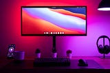 An LCD computer and peripherals on a desk. The computer displays a huge range of colors while the wall behind it is also lit in a reddish to purple spectrum — these colours often represent digital culture.