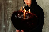 Hoodied man holding a heart-shaped candy box that’s on fire.