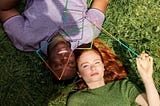 Two individuals laying on the grass with one person holding a thin colourful design sculpture