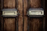 Two brass mail slots are on a wooden door. The left slot is labeled A and the right slot is labeled B.