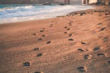 A picture of footprints in the sand with a waterbody and a bridge at the horizon, used to signify the journey and the hope that waits beyond.