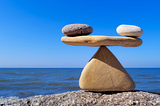 Smooth stones balanced like a scale on a rock by the ocean under a clear blue sky.