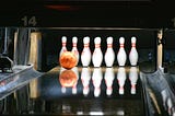 Orange bowling ball moving down an alley toward a ten pin setup.