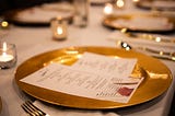 A gold dinner plate on a formal dining table with a menu in the foreground is accented by candlelight and out-of-focus dinnerware.