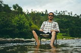 A man practising Loving Kindness Mindfulness sat in a chair in a stream outside in nature.