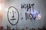 Photo of a peeling gray concrete wall. There is graffiti of a face with a neutral expression and the words “What Now?” beside it.