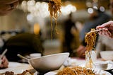 a table packed with plates and bowls of chinese food with two hands, each holding a pair of chopsticks from there are noodles hanging.