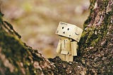 Picture of a robot looking person, made out of Amazon boxes, who is standing on a tree and looks like they are lost in the woods.