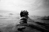 a woman, head out of the ocean, looks upward and seems to be breathing